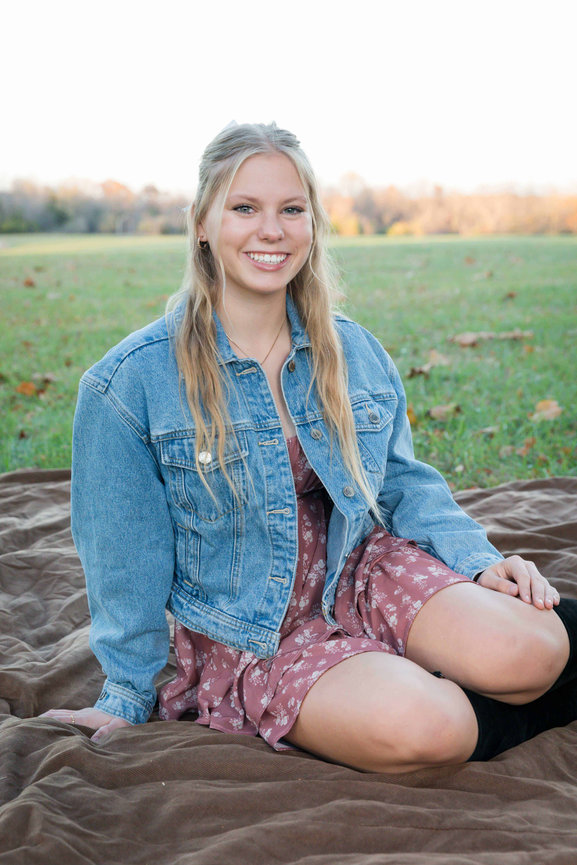 Young woman sitting on a blanket in a park, wearing a denim jacket and floral dress, smiling at the camera.