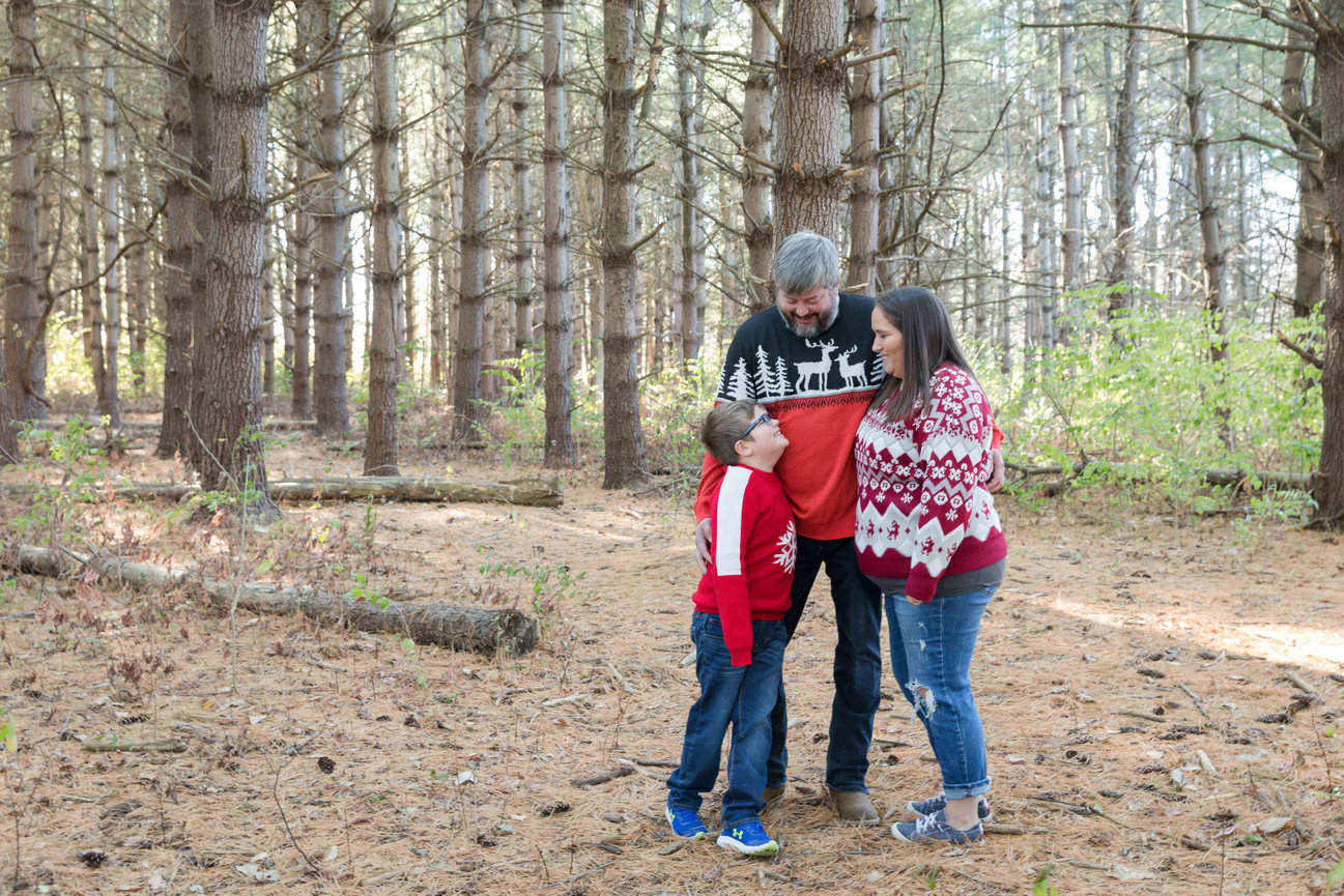 Family of three in winter sweaters standing together in a pine forest, smiling and embracing during a Blue Springs photo session photographed by Case Studios Photography