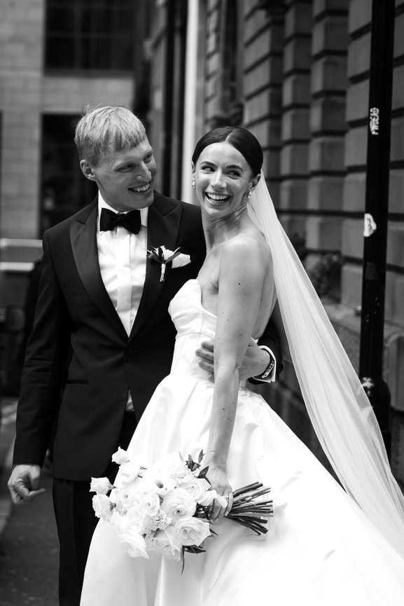 Modern city wedding photography. Bride and groom in formal attire smiling, bride holding a white rose bouquet, both standing on a Manchester city street.