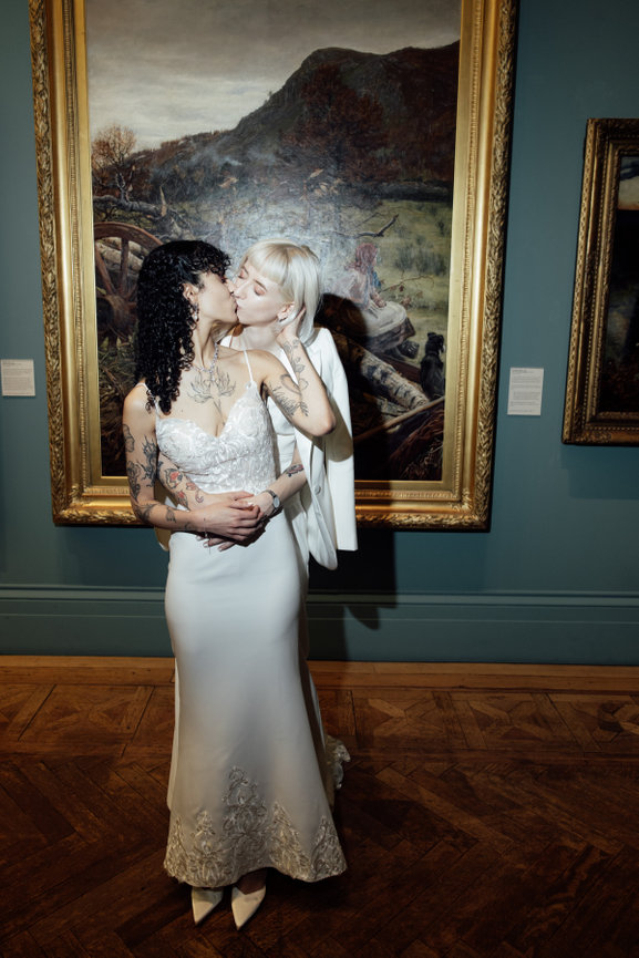 Direct flash photography: Two brides in wedding attire embrace and kiss in front of a large framed painting in Manchester Art gallery.