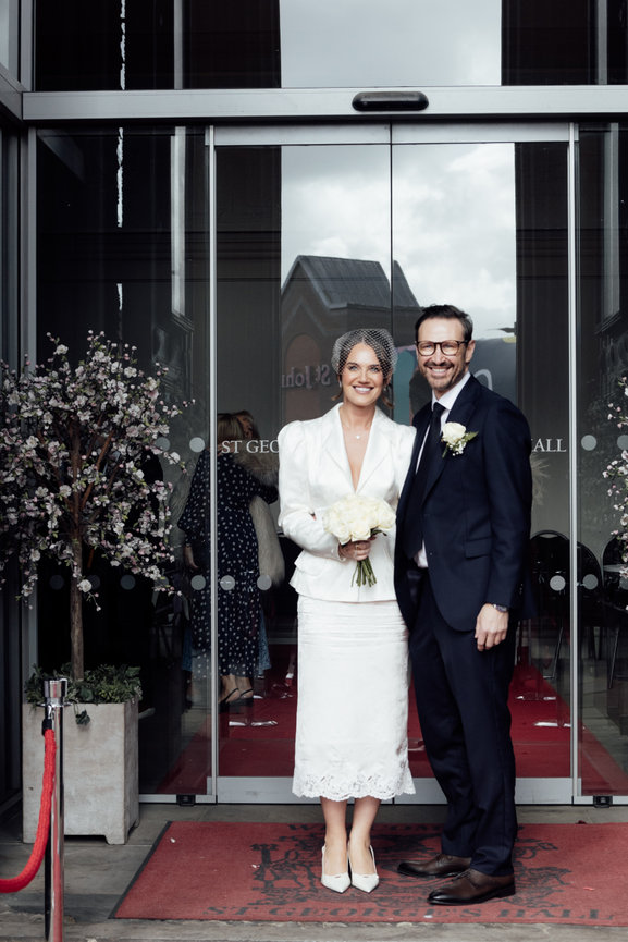 Modern micro wedding photography: A couple in formal attire smiling outside ST George's Hall, Liverpool, with floral decor and red carpet.