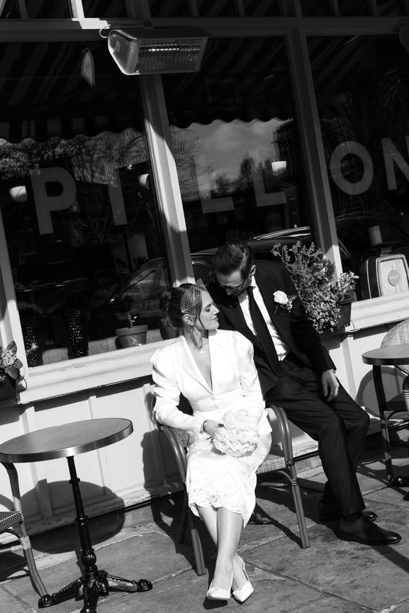 Modern editorial wedding photography of a bride and groom in vintage attire sitting outside Papillon Liverpool, captured in black and white.