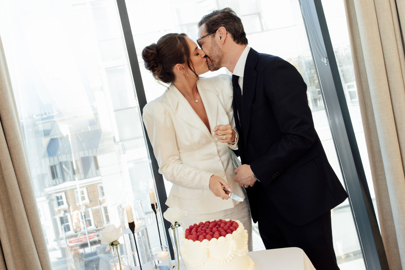Modern editorial wedding photography of a bride and groom kissing while cutting a two-tiered raspberry-topped cake at Hope Street Hotel, captured with direct flash.