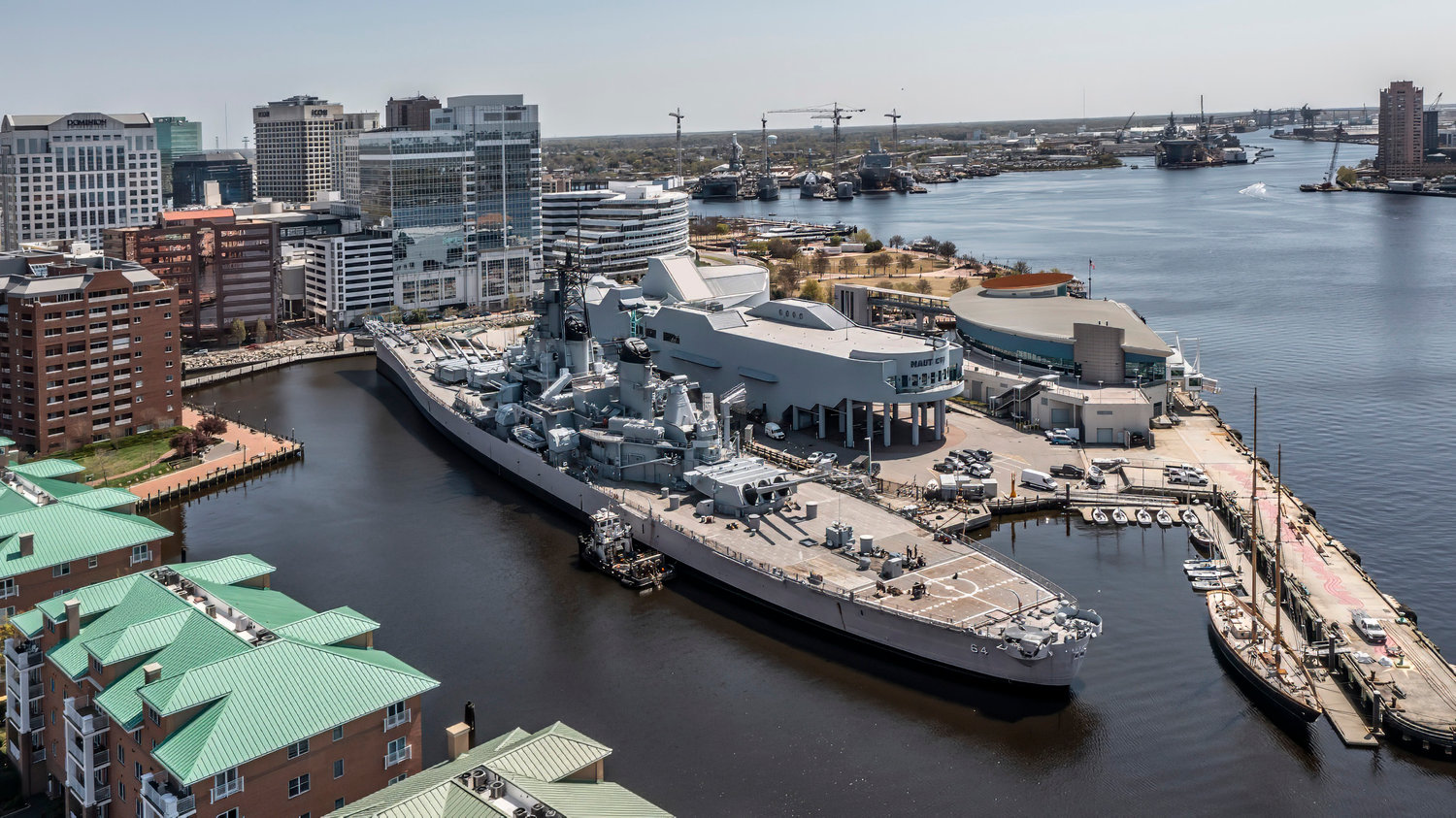 Battleship docked in a harbor cityscape with skyscrapers and cranes in the background.