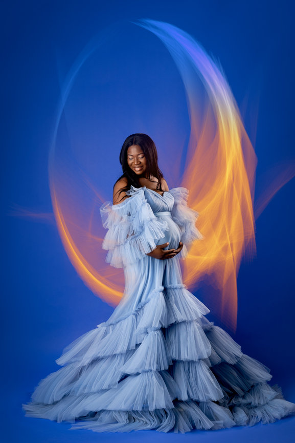 Pregnant woman in a flowing blue gown stands against a blue backdrop with colorful light swirls.