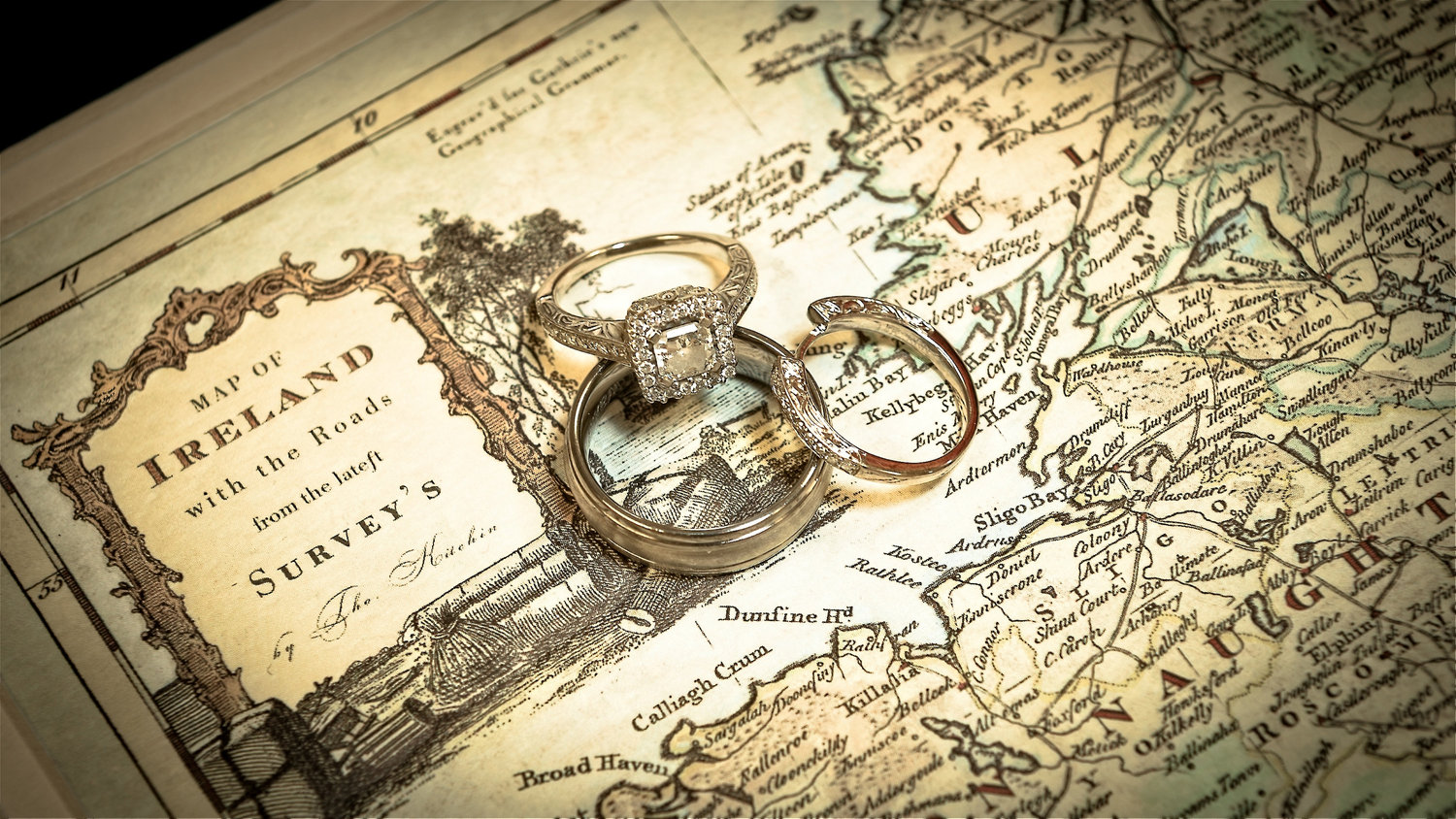 Close-up of three wedding rings, including a diamond engagement ring, resting on an antique map of Ireland, with the rings placed over the northwest coastal region.