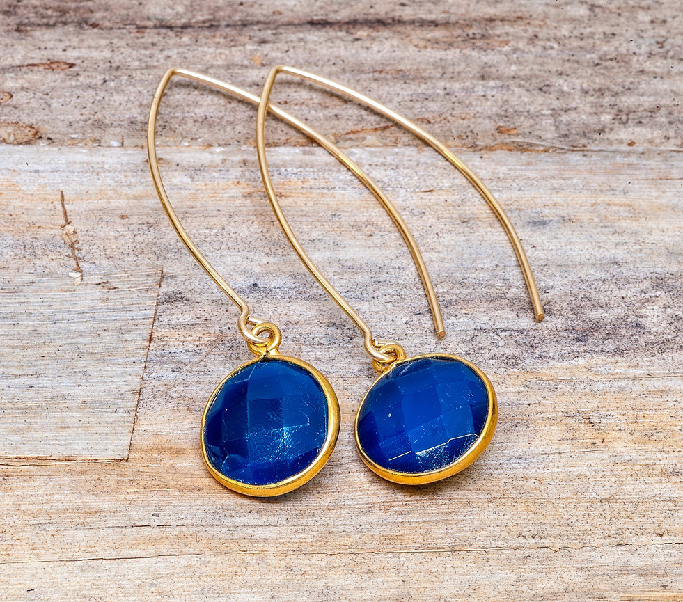 Pair of gold drop earrings with elongated hooks, featuring round faceted deep blue gemstones in gold bezels, displayed on a rustic wooden surface.