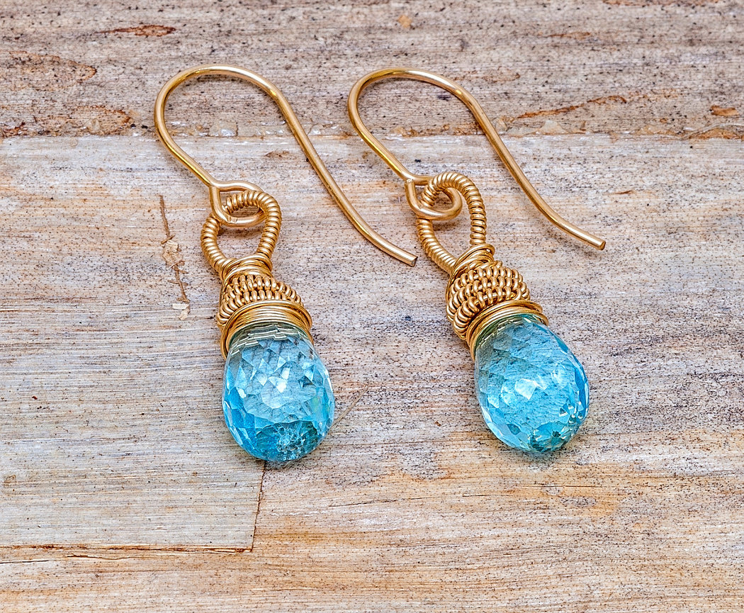 Pair of handmade dangle earrings featuring faceted teardrop-shaped blue gemstones wrapped in intricate gold wire, attached to gold hook ear wires, displayed on a rustic wooden surface