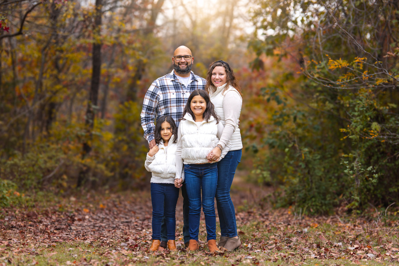Family of four standing together in a forest with autumn foliage, smiling and wearing cozy outfits in Richardson, Texas