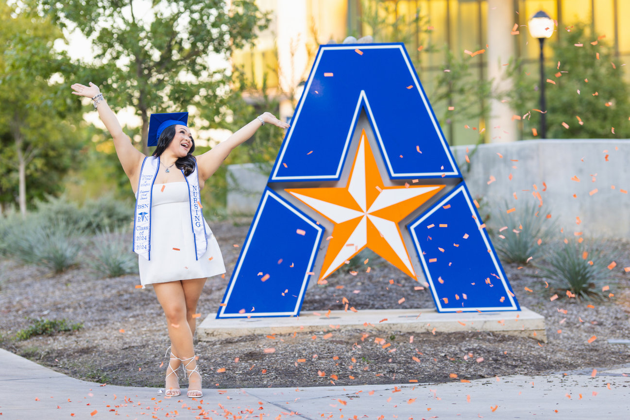 Graduate celebrating in front of a large blue and orange A symbol at the University of Texas at Arlington with confetti falling around her.