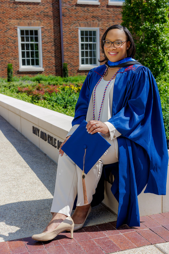 Graduate in blue gown and tassel, sitting outdoors on Southern Methodist in Dallas, Texas, campus with diploma, smiling confidently.