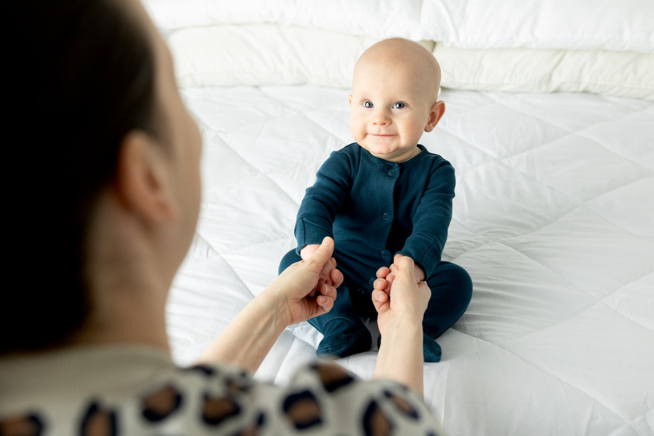 A smiling baby in a blue onesie sits on a bed, holding hands with an adult.