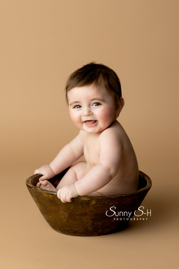 Smiling baby sitting in a wooden bowl against a plain background.
