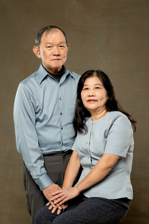 Elderly couple posing together against a neutral background, both wearing light blue shirts.