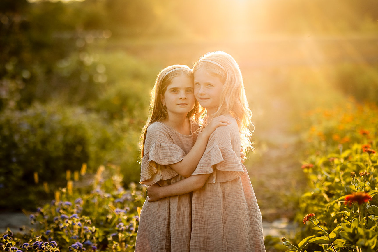 Two young girls in light dresses stand in a sunlit garden surrounded by colorful flowers, gazing at the camera.