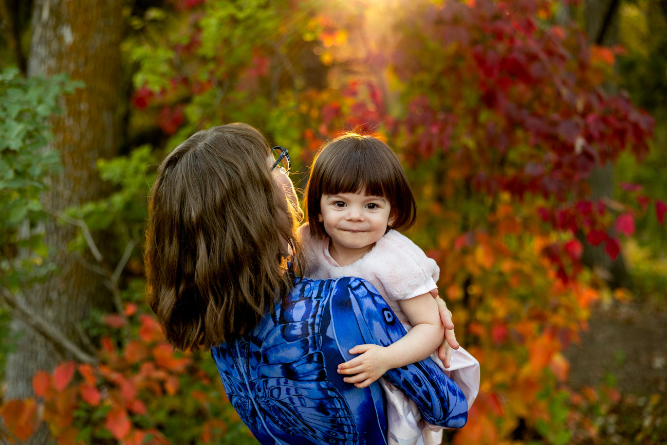 Adult holding a smiling child outdoors, surrounded by vibrant autumn leaves and sunlight filtering through trees.