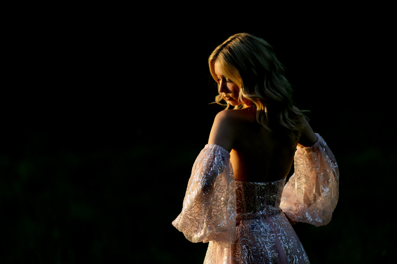 Back view of a woman in a sheer embroidered dress against a black background, lit by warm side light