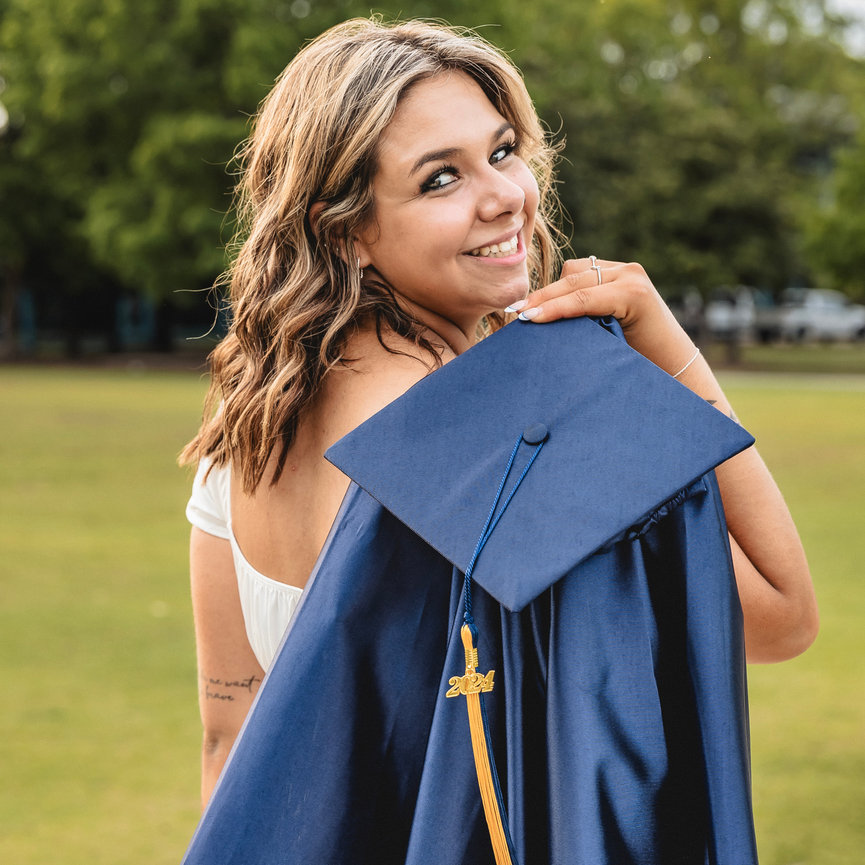 Smiling graduate holding a blue cap and gown outdoors on a grassy field.