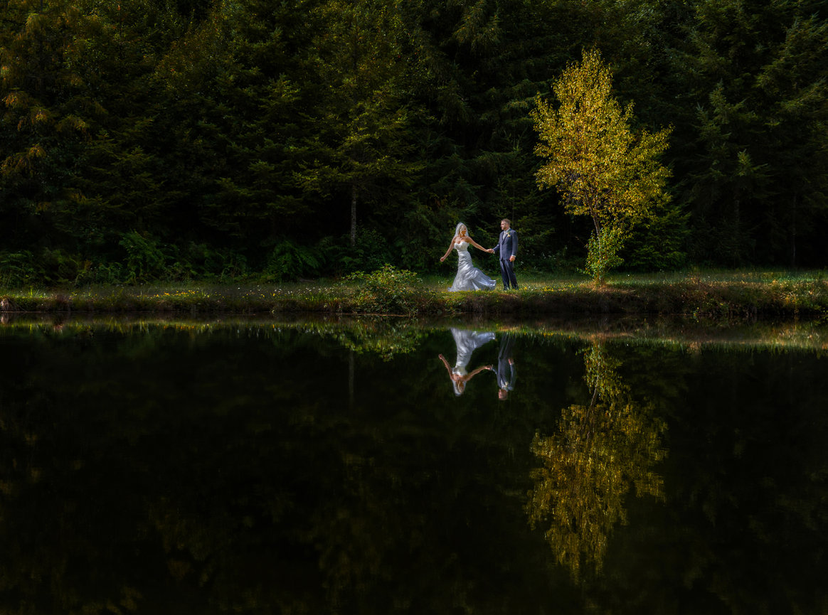 Couple standing by a calm lake with a clear reflection of the trees.