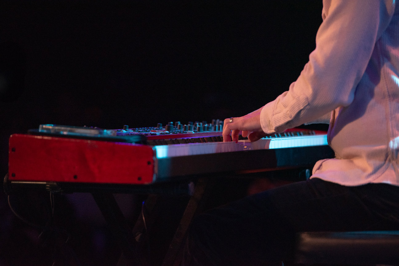 A close-up of a musician's hands playing a keyboard on stage, with vibrant lighting in the background.