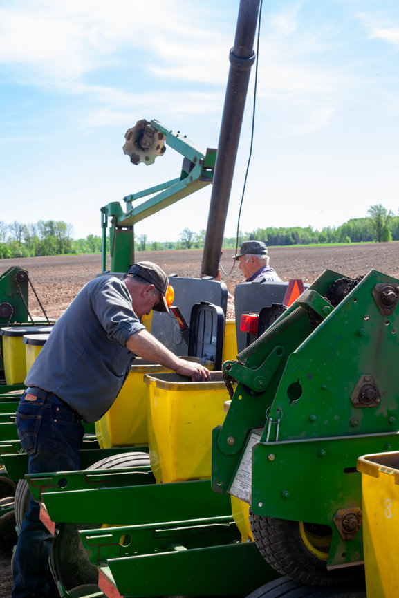 Farmers loading yellow seed boxes into a green planter in a freshly prepared field during spring planting under a bright blue sky