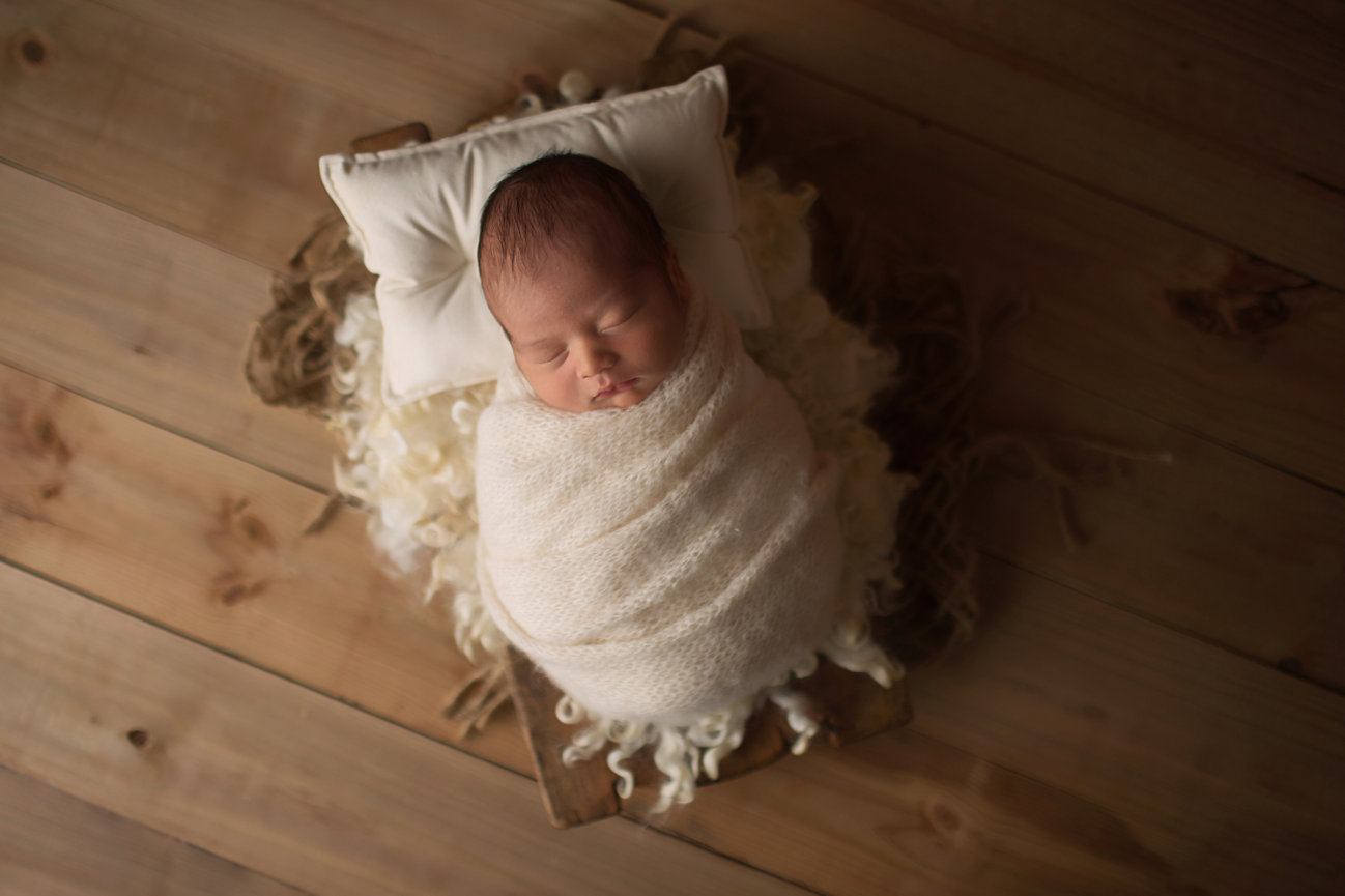 Newborn baby wrapped in cream knit wrap laying on cream velvet pillow and cream curly fur in a wooden trench bowl with wood background with Kansas City photographer Dreamworks Photography