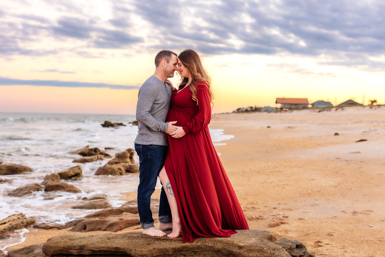 Couple embracing on a rocky beach at sunset, with the woman in a long red dress during a maternity photography session with Catherine Whitney Photography in Jacksonville Florida