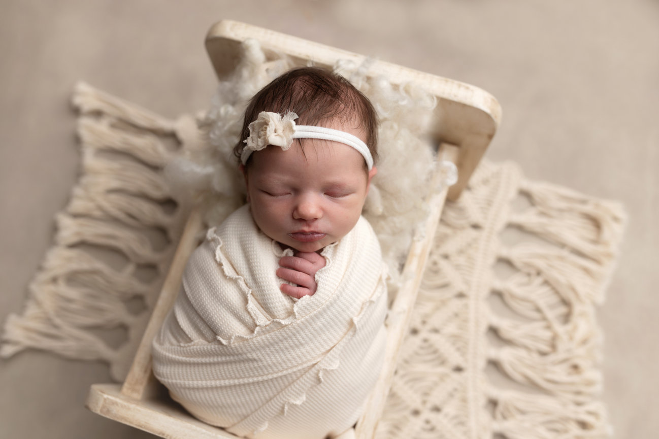 Newborn baby sleeping in a small, white wooden bed, wrapped in a cream blanket, wearing a headband with a flower.