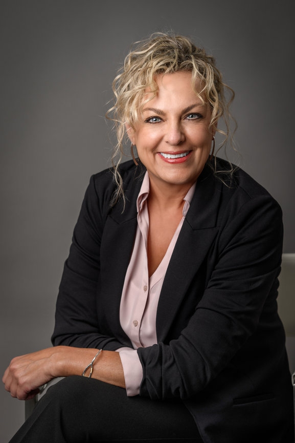 Professional portrait of a smiling woman with curly blonde hair, wearing a black blazer over a pink blouse.