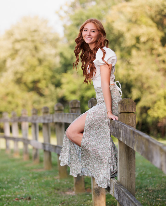A beautiful high school senior red head girl sits on a wood fence on a country road in Carmel Indiana