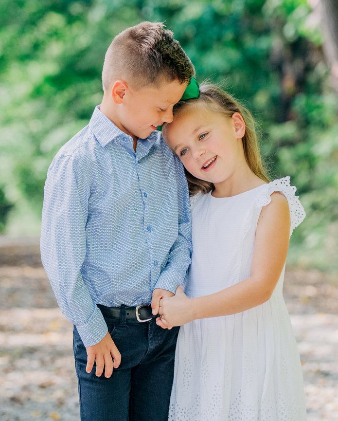 A cute brother dressed in a blue button down and his sister in a white dress snuggle with each other on a country road in a wooded area in Fishers Indiana.