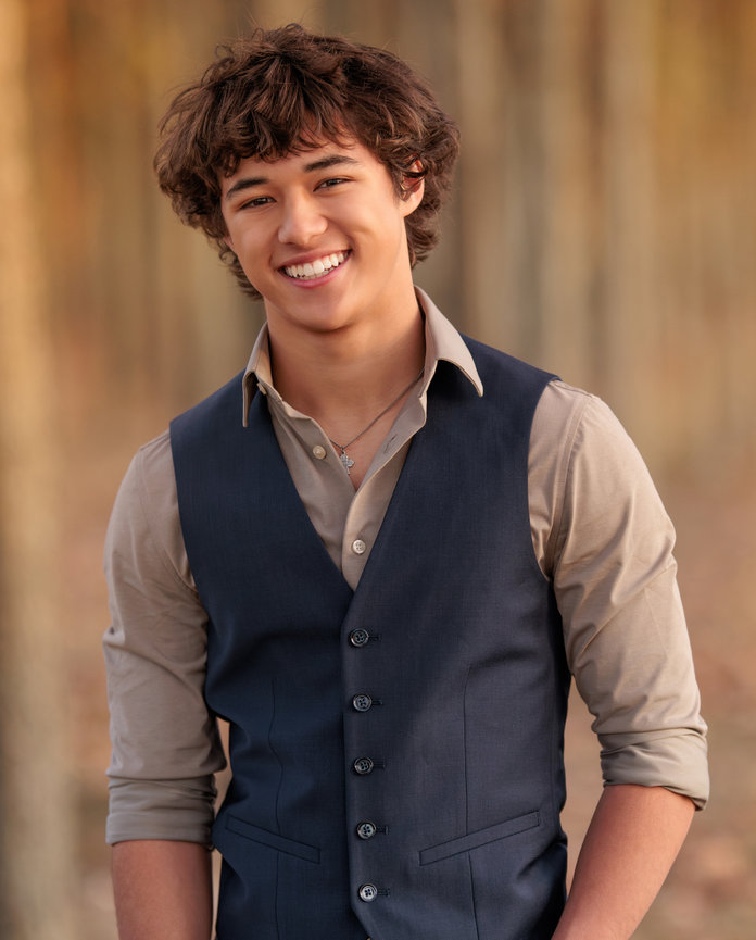 A good looking brunette high school senior boy wearing a blue vest smiles while standing in a forest in the fall in Indianapolis Indiana.