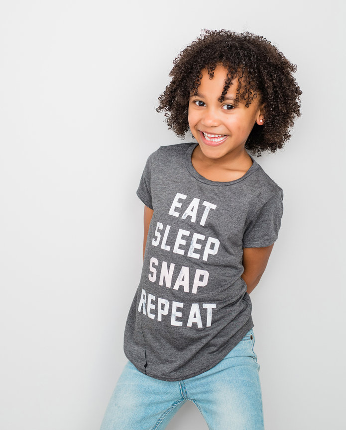 Young girl smiling, wearing a gray shirt with EAT SLEEP SNAP REPEAT text and blue jeans, standing against a plain white background.
