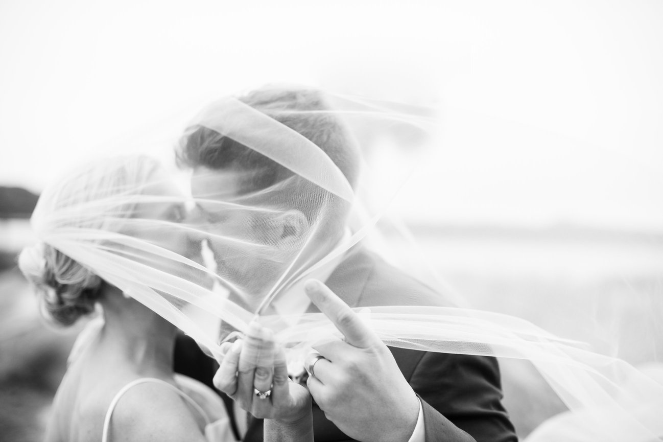 Black and white image of veil blowing as bride and groom kiss by the marsh in South Carolina. South Carolina destination wedding photography by Heather Durham.