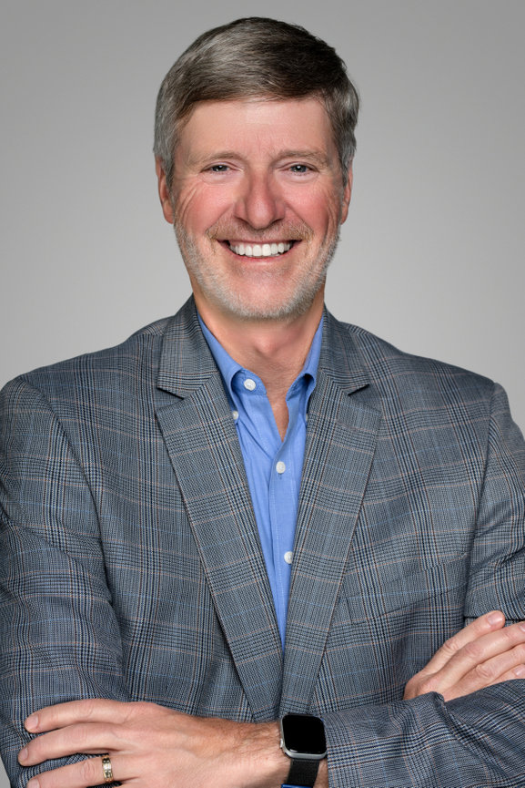 Middle-aged man in a gray plaid blazer, blue shirt, smiling with arms crossed against a neutral background.