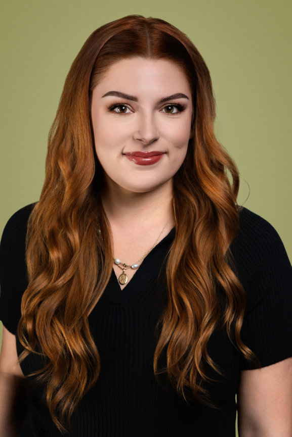 Woman with long, wavy red hair wearing a black top, smiling against a green background.