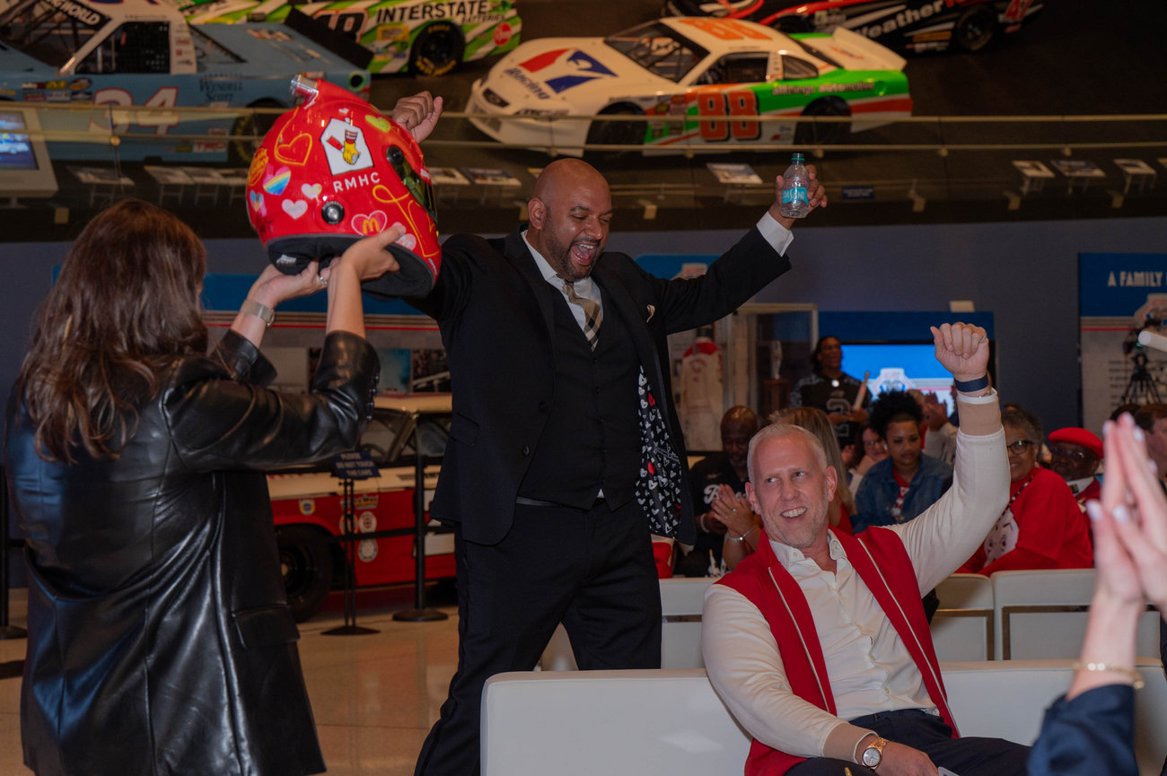 People celebrate indoors near race cars. One person holds a helmet. Audience members clap and cheer captured in the style of Jo Hayes Images Charlotte Event Photography Event Coverage