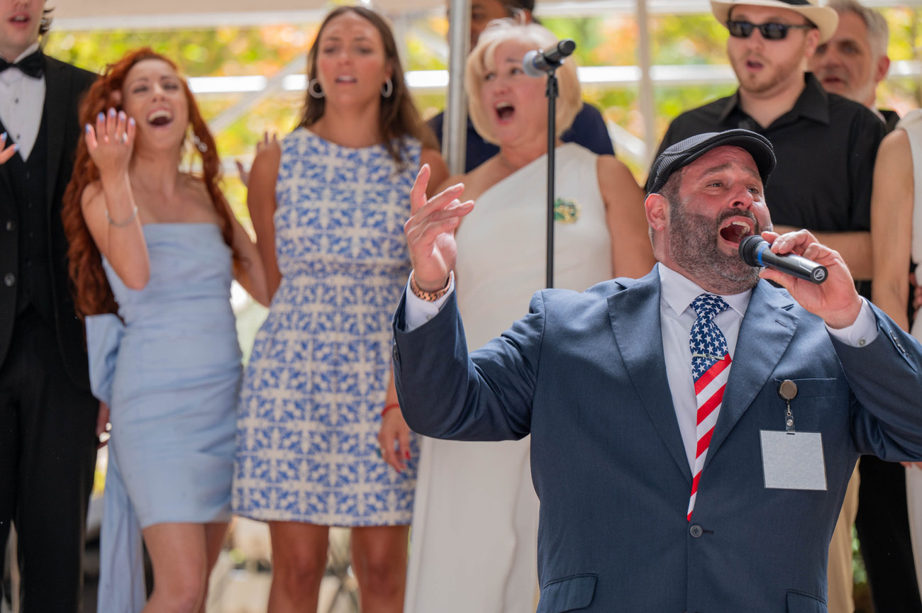 A group of people, dressed formally, sing while a man in a suit and cap performs with a microphone at Festa Italiana Uptown Charlotte captured by professional photographer Jo Hayes Images Charlotte Event Photography