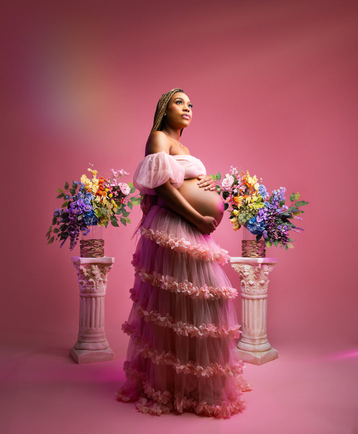 Pregnant woman in a pink floral dress posing against a pink backdrop with colorful flower arrangements during a Baltimore maternity photography session