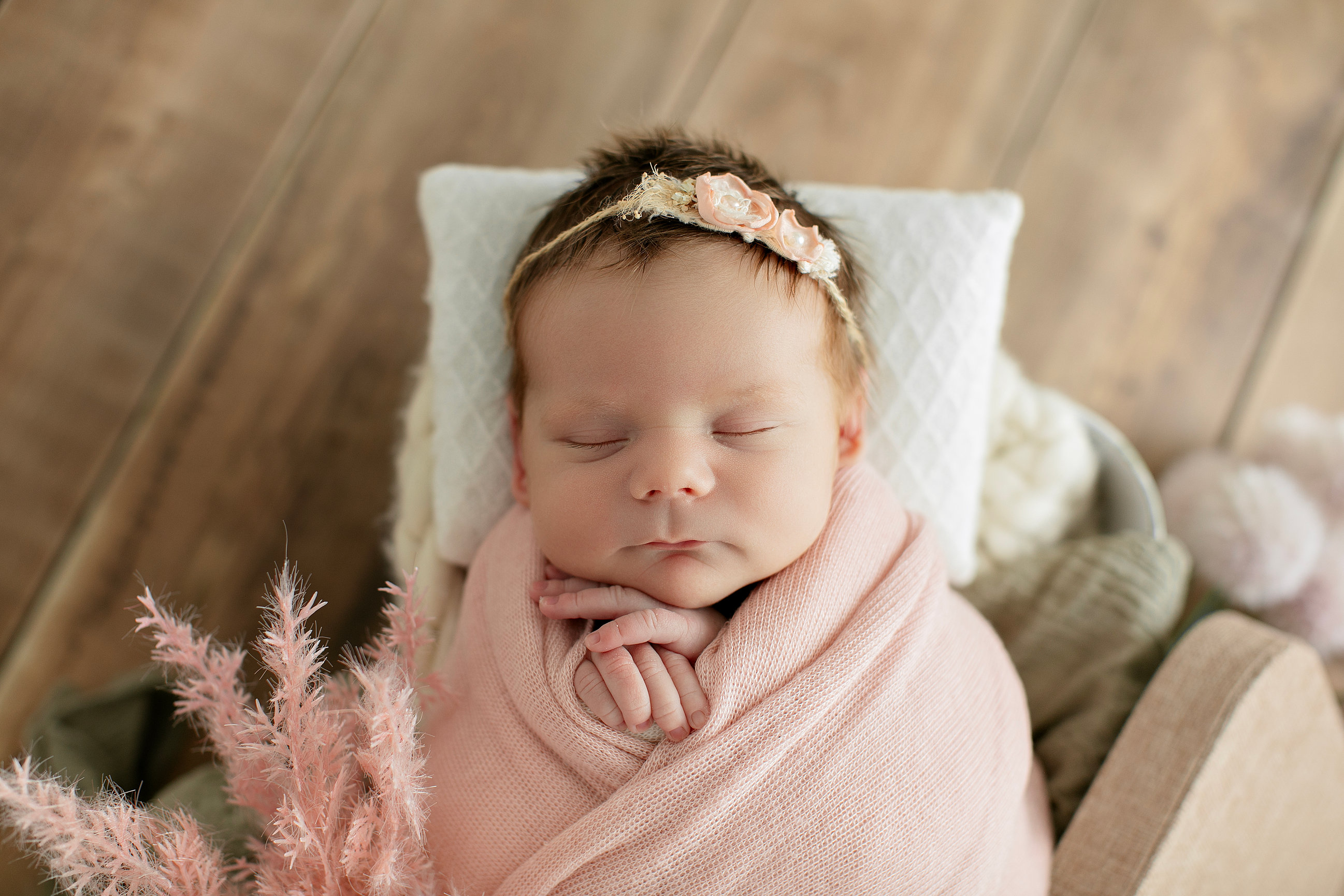 Sleeping newborn wrapped in pink, resting on a tiny pillow beside dried flowers.
