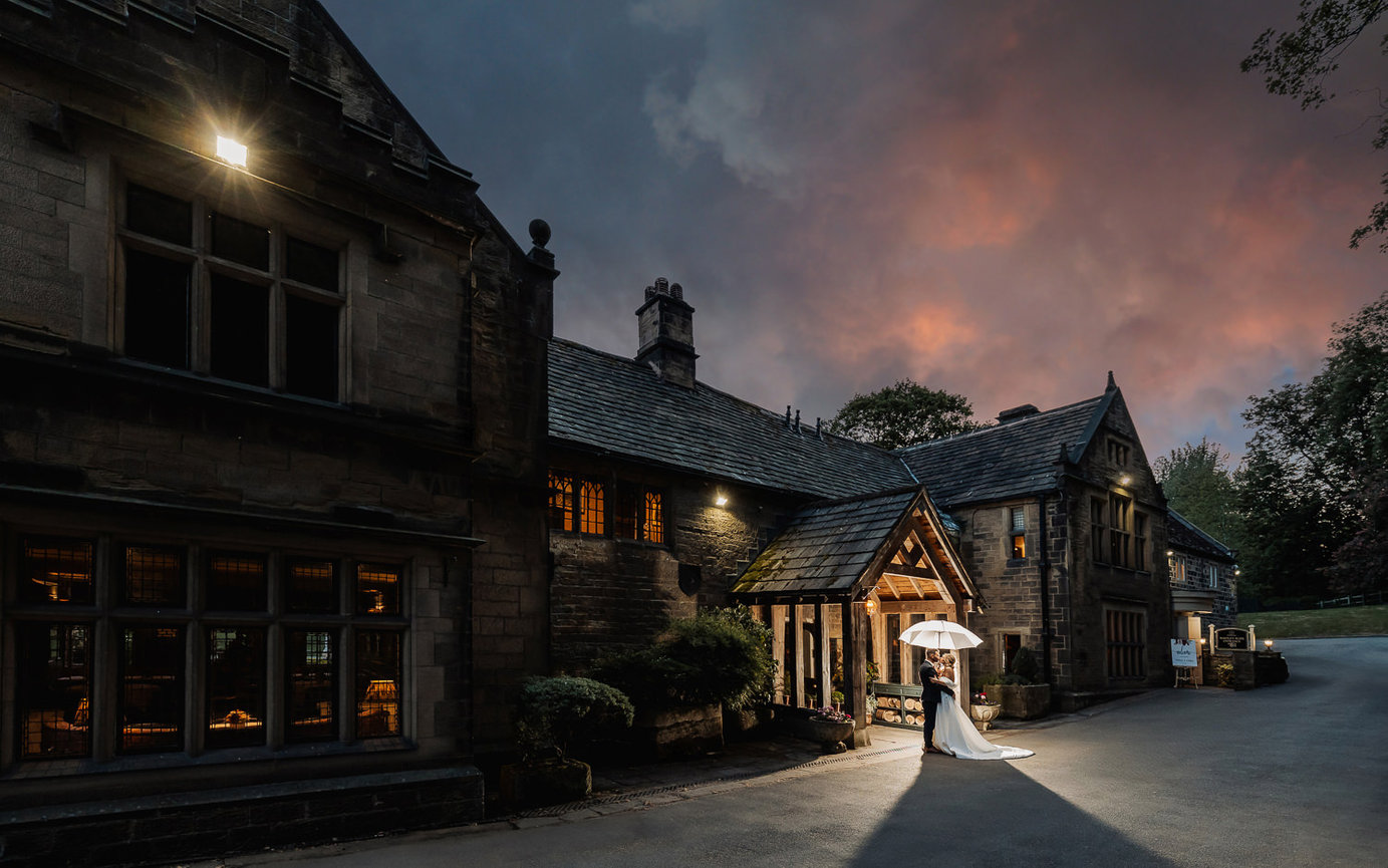 Bride and groom under umbrella by a historic stone building at dusk, with a dramatic sky in the background.