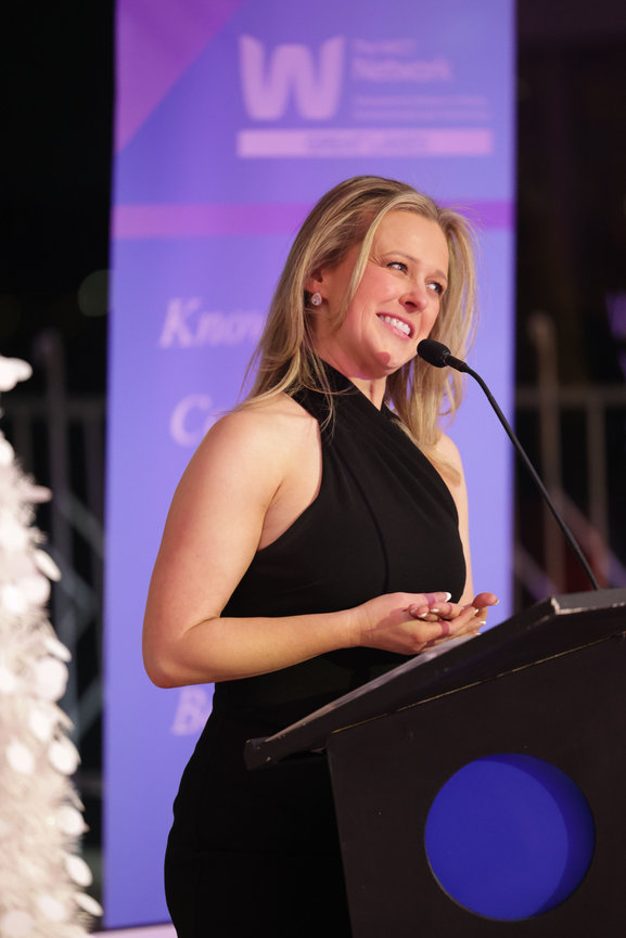 Woman in a black dress speaks at a podium with a microphone, in front of a blue and purple backdrop.