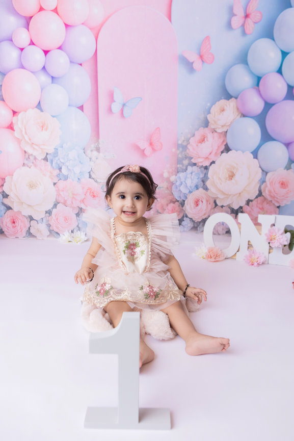 Smiling baby in a pink dress with a pastel floral backdrop and white ONE letters