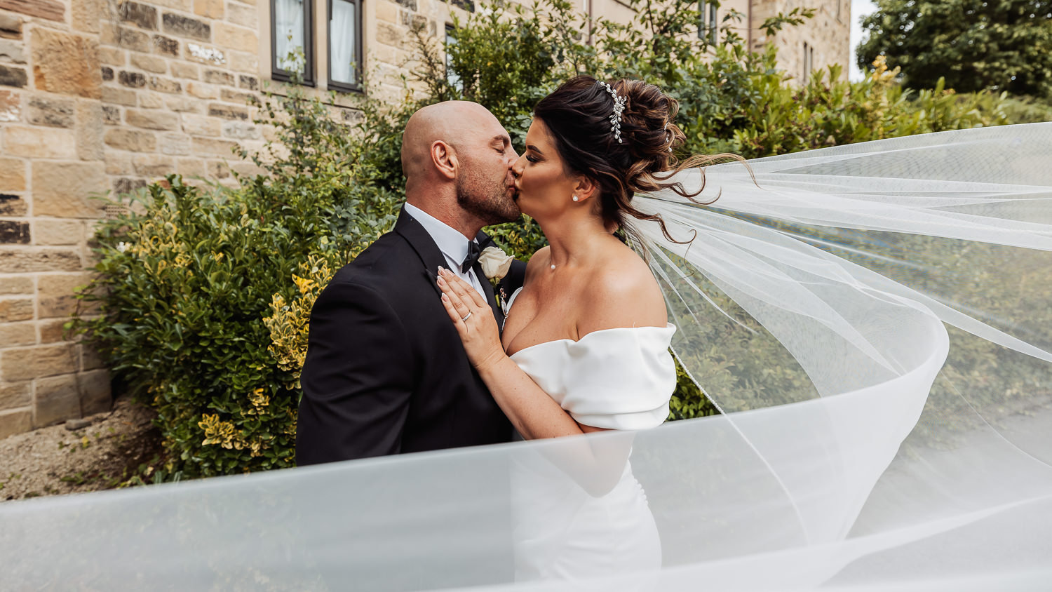 Bride and groom kissing on a curved road, with a flowing veil and stone building in the background