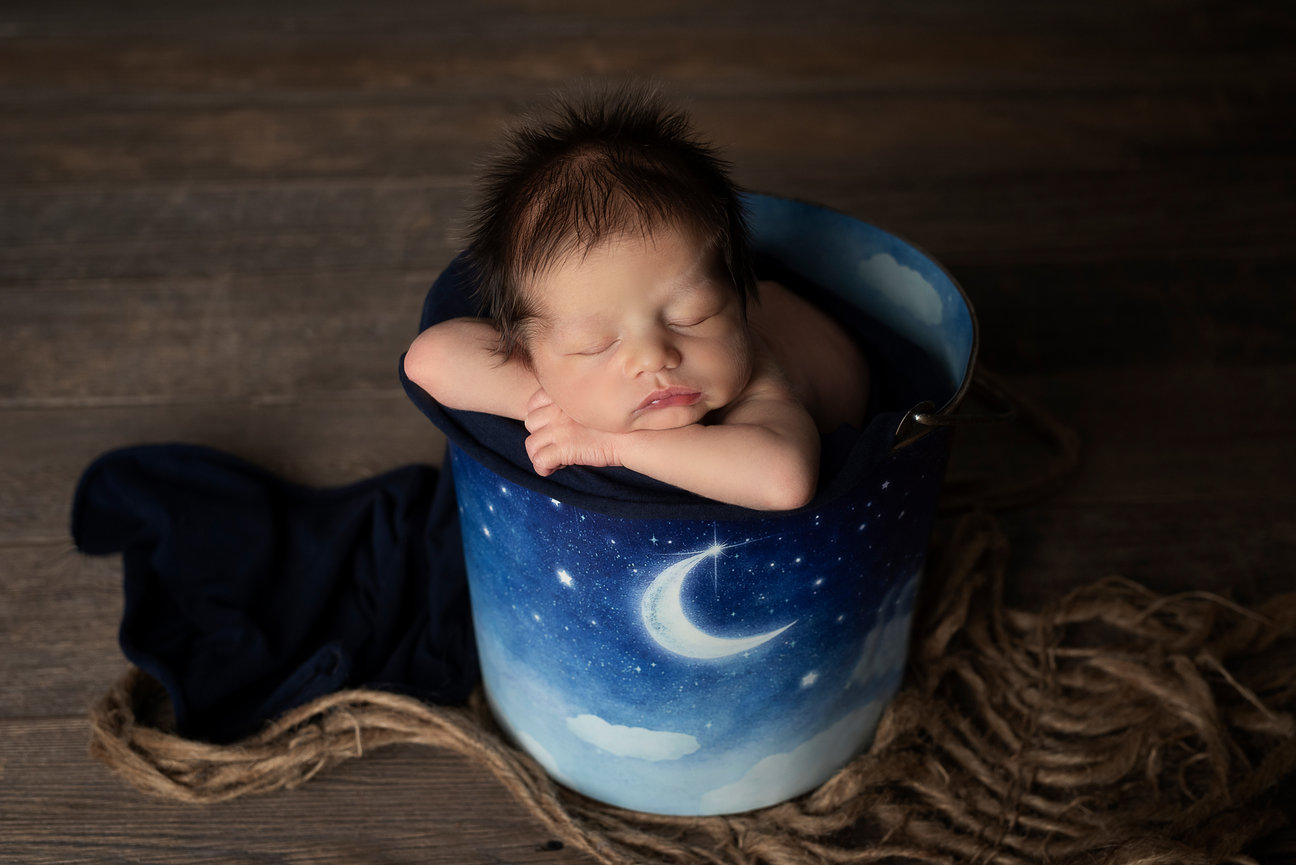 A newborn baby sleeps peacefully with arms folded, nestled in a blue bucket adorned with a crescent moon and stars pattern. This charming scene unfolds on a wooden floor in a cozy Streamwood home, where dark cloth and textured fabric.