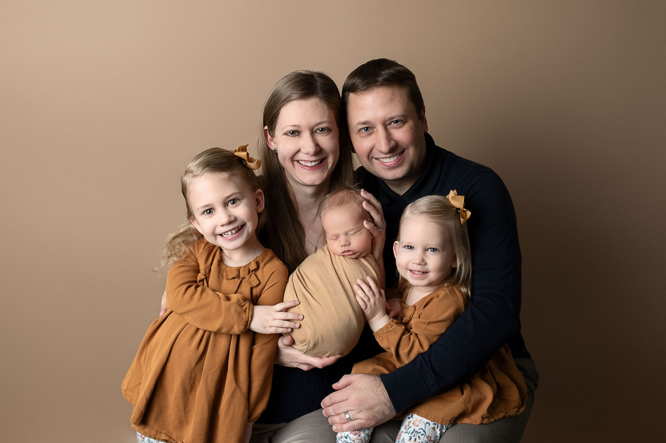 Family portrait with two adults and three children in Hampshire. The mother lovingly holds a newborn wrapped in tan fabric, while the father embraces the group. Two young girls in matching brown dresses sit beside them, all smiling.