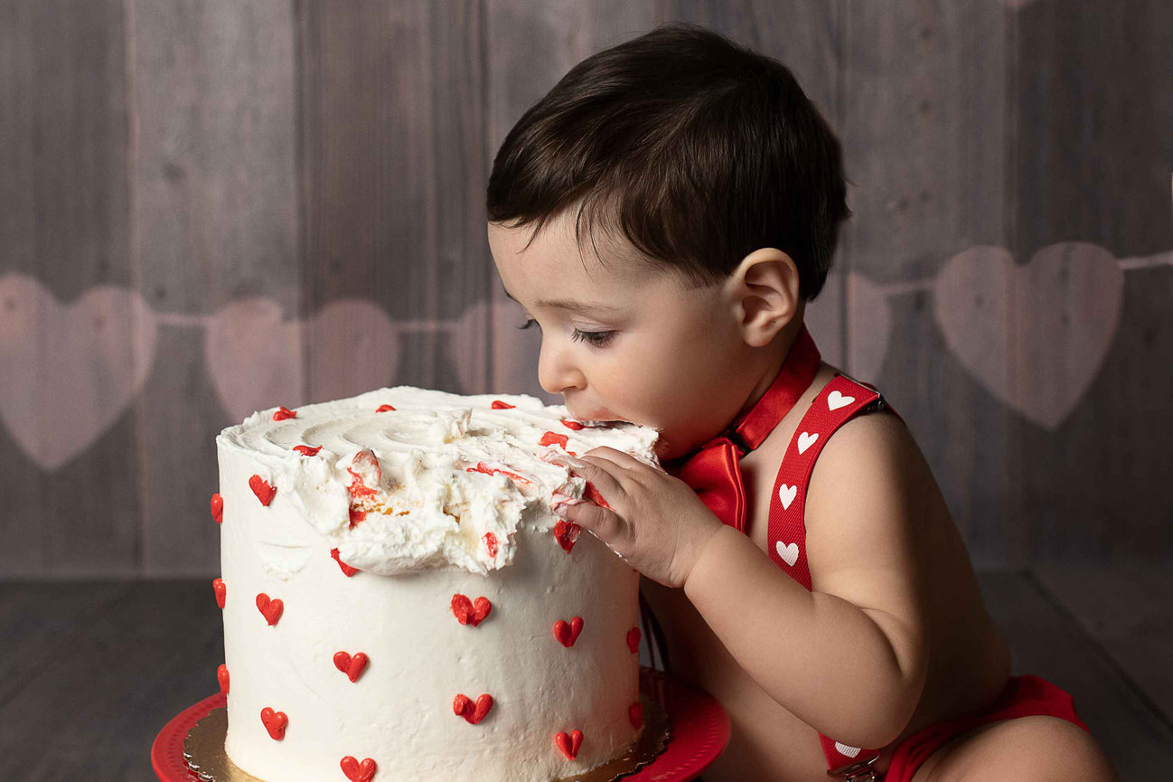 A toddler with dark hair wearing a red outfit adorned with white hearts is eagerly biting into a tall, white cake covered in red heart-shaped sprinkles. Against the backdrop of Chicagolands signature wooden textures and faint heart-shaped decorations