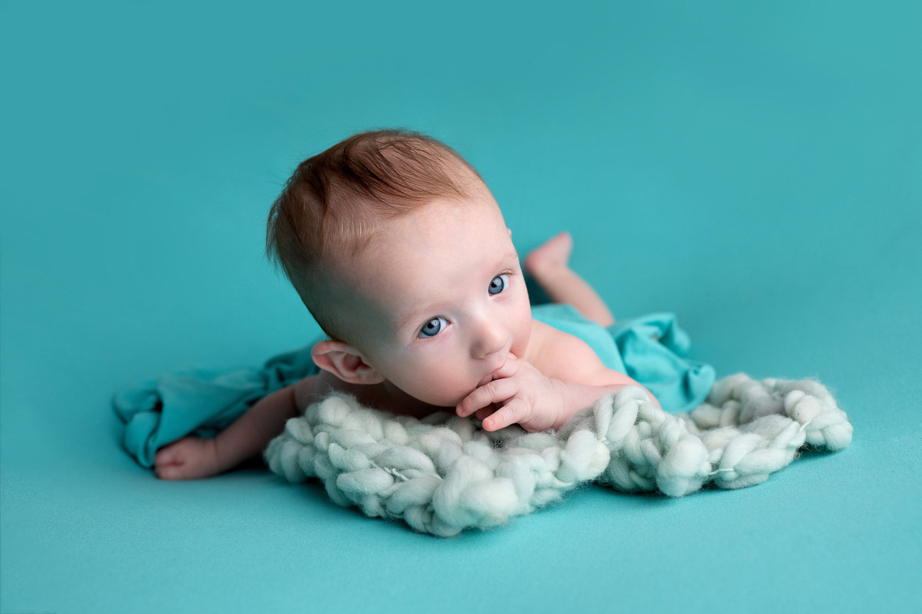 A baby lies on their stomach on a soft turquoise blanket, surrounded by light, fluffy white fabric. The little one gazes upwards with their thumb in their mouth, perfectly capturing the essence of baby photography against a turquoise background.