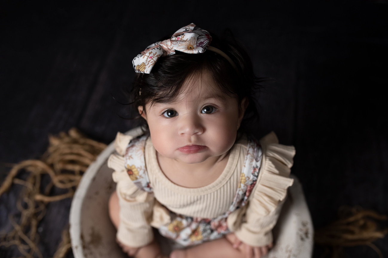 A baby with dark hair and big eyes sits in a bowl on a dark floor, embodying the charm of children photography. They wear a floral outfit with ruffled sleeves and a matching headband with a bow. Rug strands peek beside the bowl.