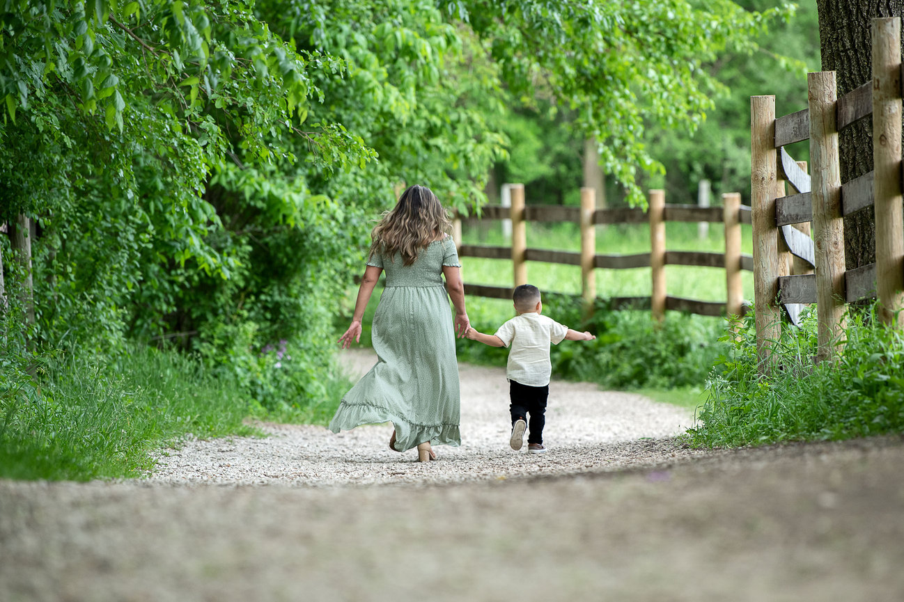 A woman in a green dress walks hand in hand with a child down a gravel path lined with a wooden fence and lush green trees, as if captured by Carol Stream Photography, suggesting a peaceful, rural scene.